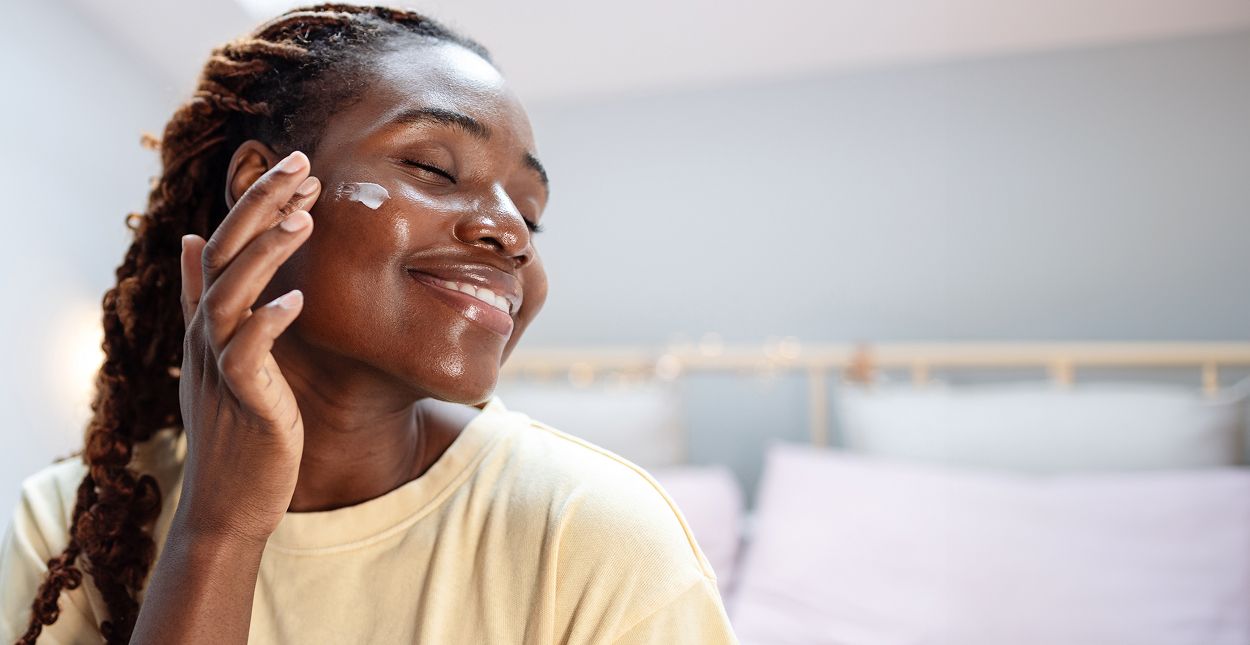 Woman applying skincare cream, smiling happily.